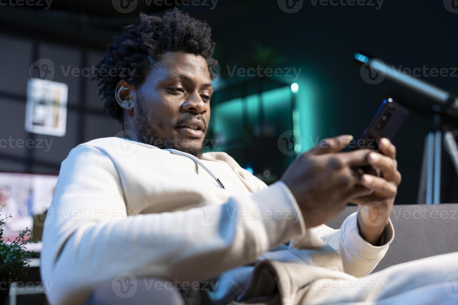 Joyous man relaxing by texting friend, discussing meeting hour. BIPOC person sitting on cozy sofa in modern stylish living room typing on mobile phone, excited to meet up with mate photo