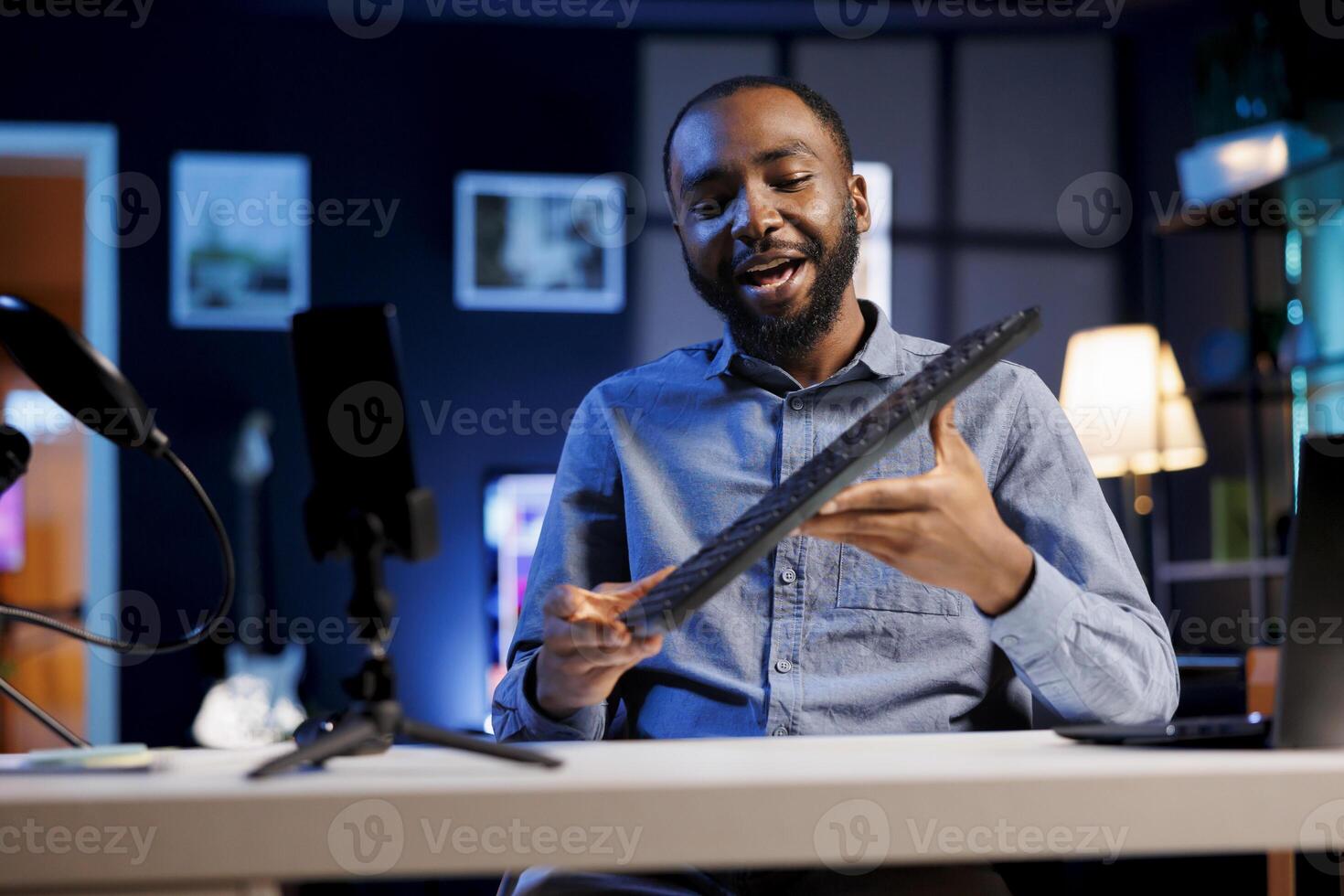 African american internet show host using smartphone camera on phone holder to film content about best computer peripherals of the year. Online star recording mechanical keyboard review photo