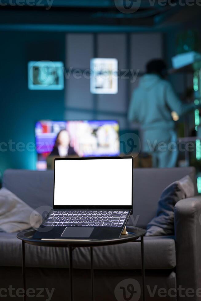 Focus on isolated screen laptop on coffee table in RGB lit home at night with man in blurry background looking on shelves. Close up shot of mockup notebook and person in the back doing inventory photo