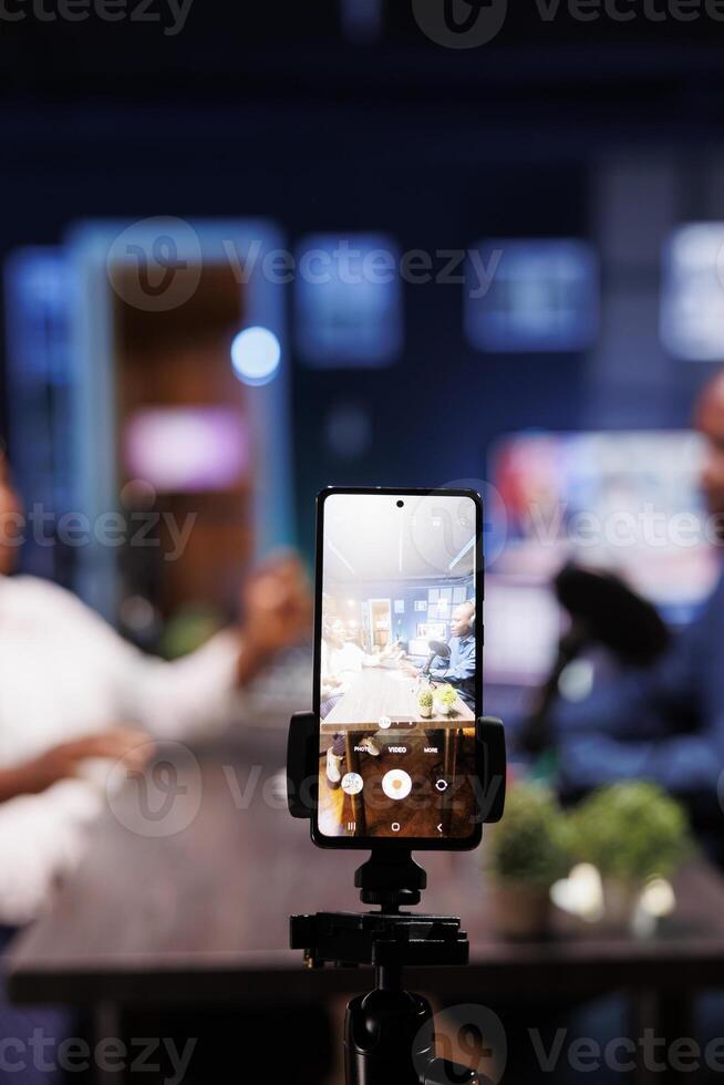 In a neon-lit studio, a vlogger show host is interviewing a guest while a smartphone on a tripod records them. The camera is focused on the phone capturing the content of the influencers. photo