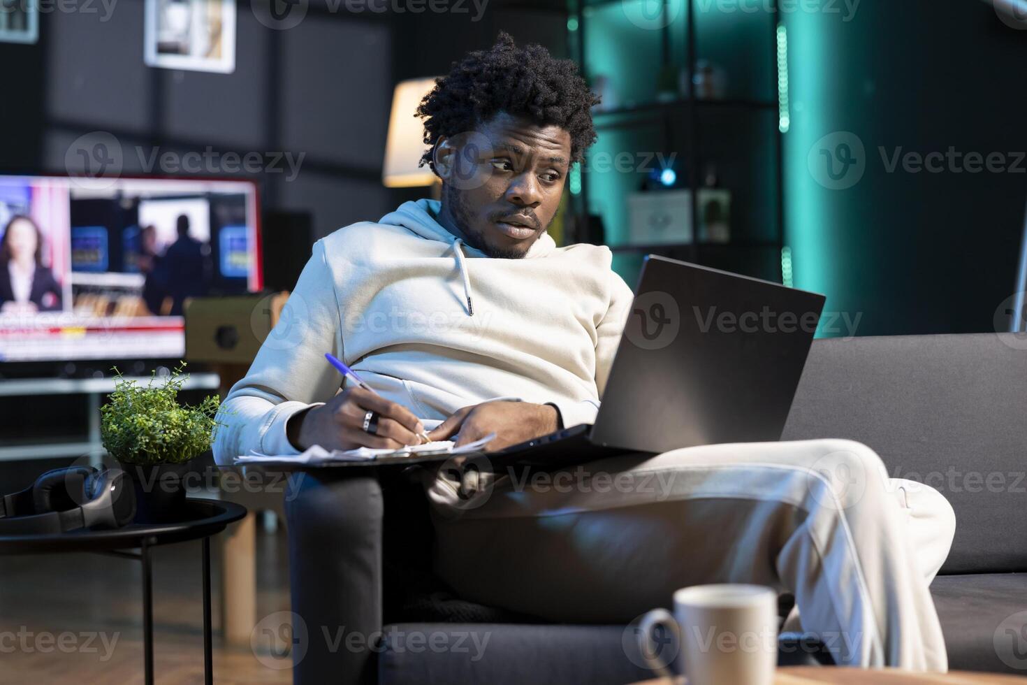 Self employed man working from home, writing business ideas, looking at laptop screen. BIPOC man sitting on couch with notebook in lap, using pen to note plan on paper while teleworking in apartment photo