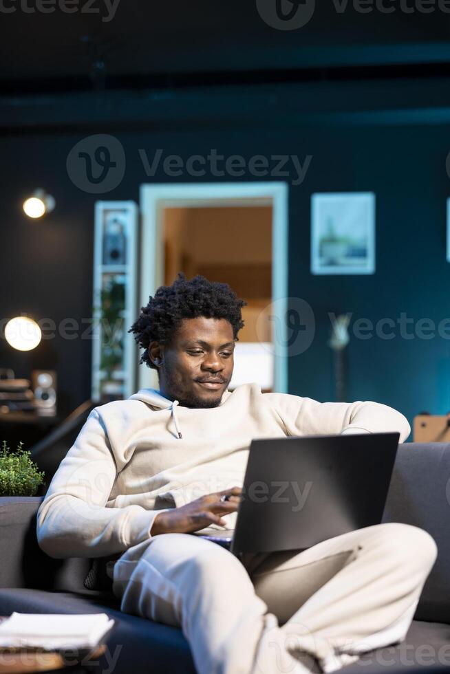 Smiling self employed man checking his emails on laptop while laying on couch. Cheerful remote employee working from home in bright stylish cozy apartment personal office, reading news on notebook photo