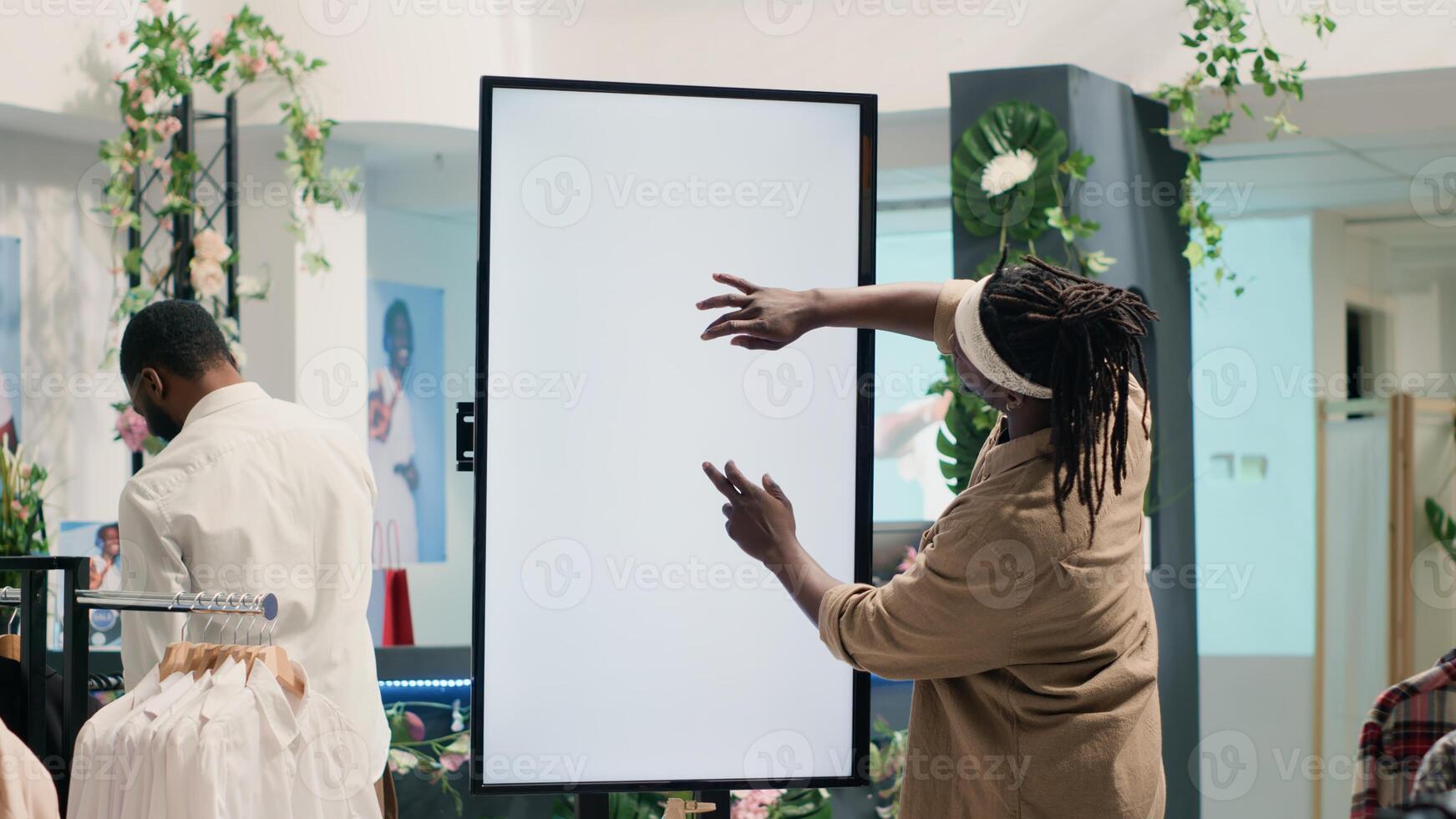 BIPOC client in clothing store using augmented reality mockup screen to look at clothes options to try on. Customer using led kiosk to visualize outfits in showroom before buying photo
