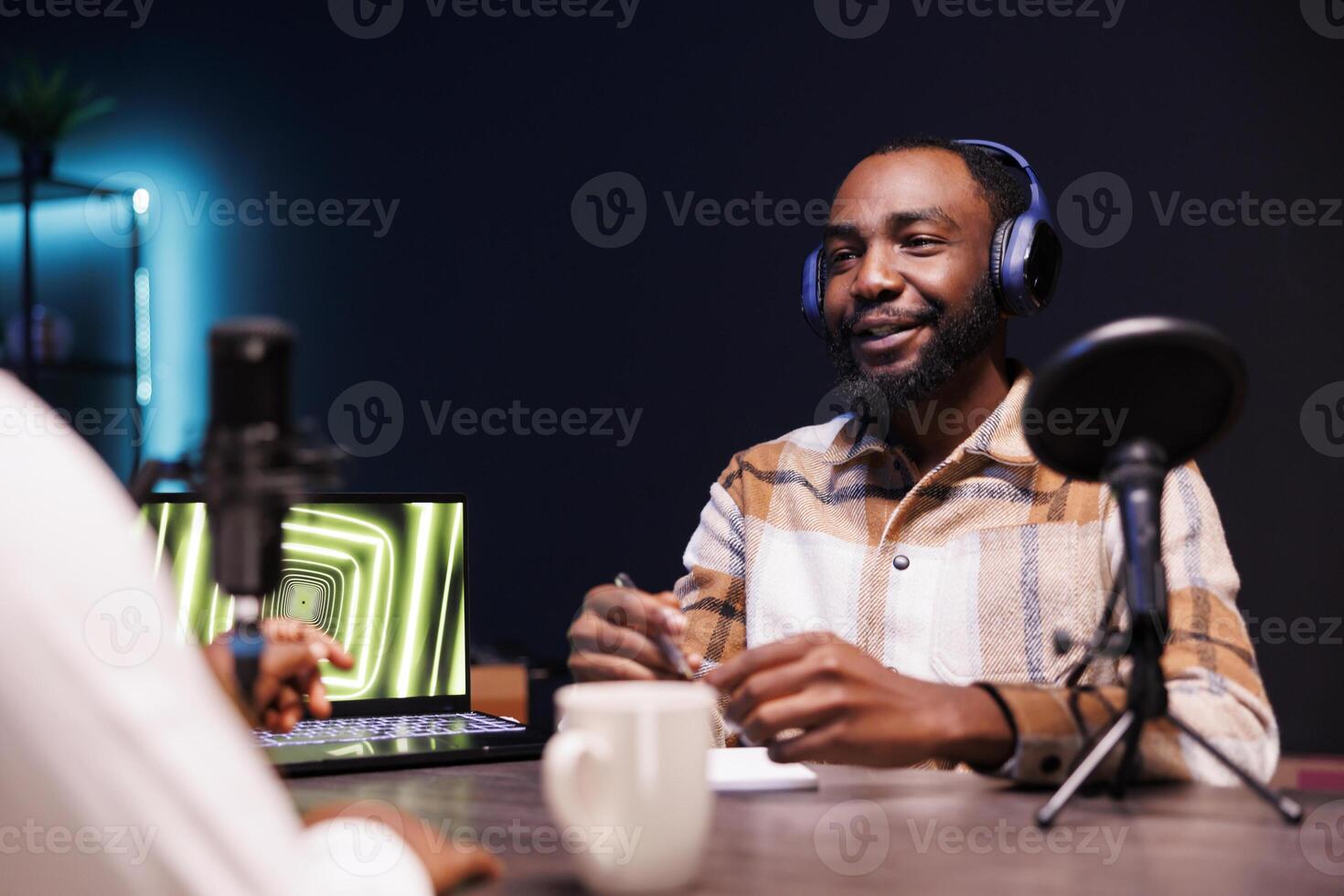 Black guy with a notebook is using podcast equipment to provide online advice. Male African American vlogger is seated at the table, recording an episode at home while speaking into a microphone. photo