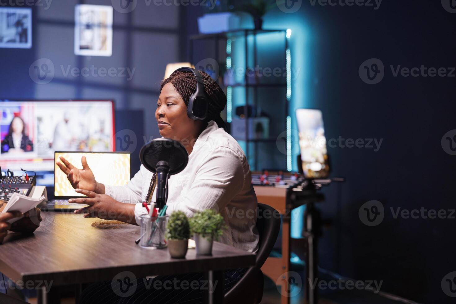 Female celebrity is seen answering interviewer questions in home studio, utilizing smartphone on tripod to record encounter. Black woman filming content for online audience, sharing exciting stories. photo
