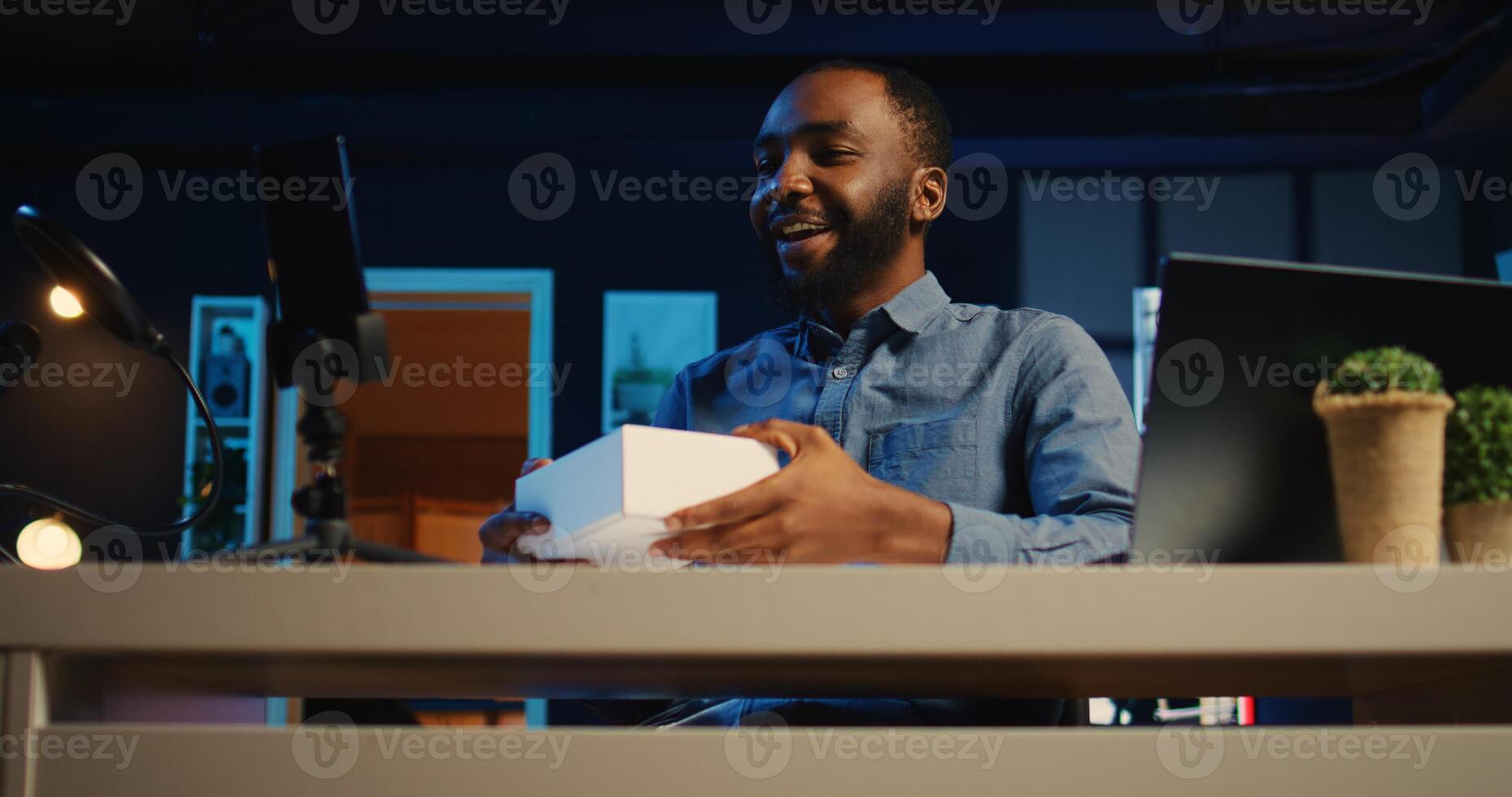 Tech content creator filming packaged product unboxing in RGB lights personal home studio. Influencer showing sponsoring branded merchandise to subscribers, hip level shot photo