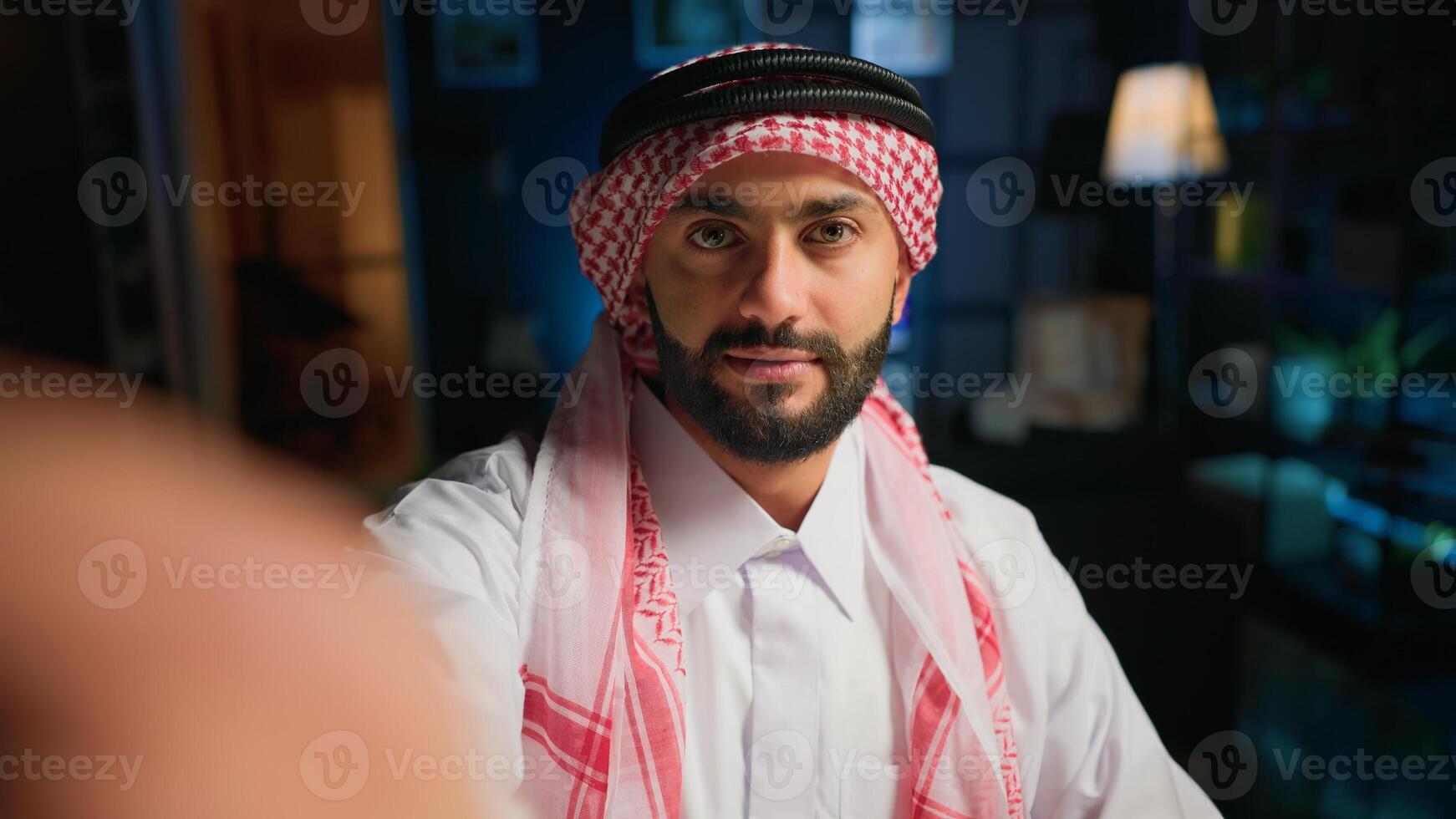 POV of cheerful Muslim content creator dressed in traditional clothing filming himself. Smiling Arab man with thobe and headscarf waving towards camera, doing online vlogging photo