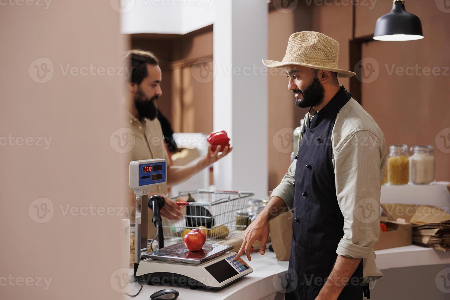 Caucasian man buying fresh produce while vendor uses electronic scale at organic supermarket checkout. Storekeeper with a hat is weighing organic fruits and vegetables while customer waits. photo