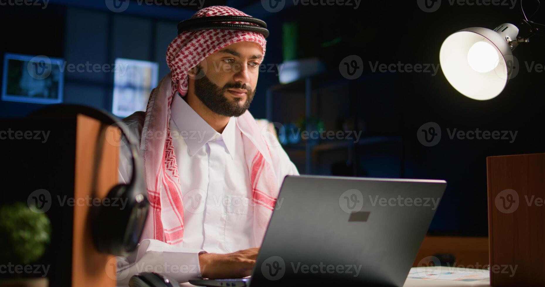 Arabic teleworker remotely working in dimly lit apartment sending emails on laptop. Middle Eastern worker at home typing business data on notebook device, wearing traditional attire photo