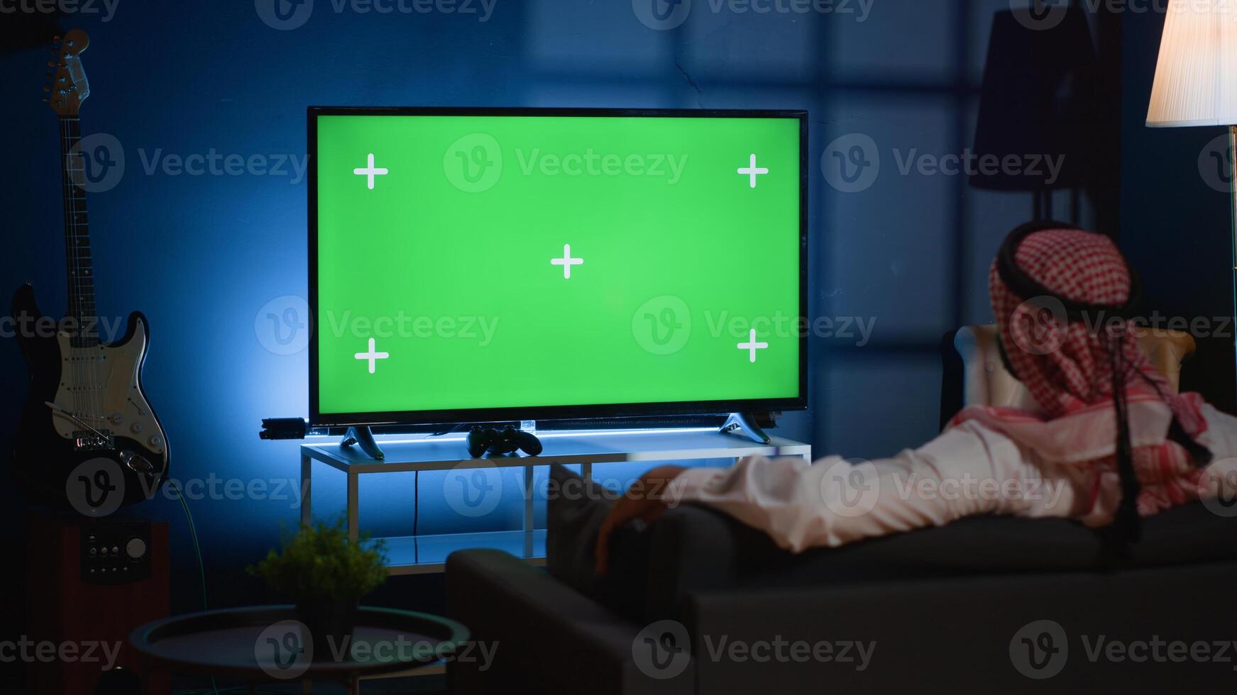Arab man lounged on cozy sofa, watching news broadcast on green screen device while relaxing at home. Muslim person chilling on couch, looking at mockup TV channel showing current events photo