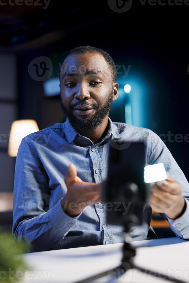 Smartphone on holder capturing footage of reviewer discussing with viewers about mini led lights. Influencer chats with fans during stream, filming RGB lighting device with phone camera photo