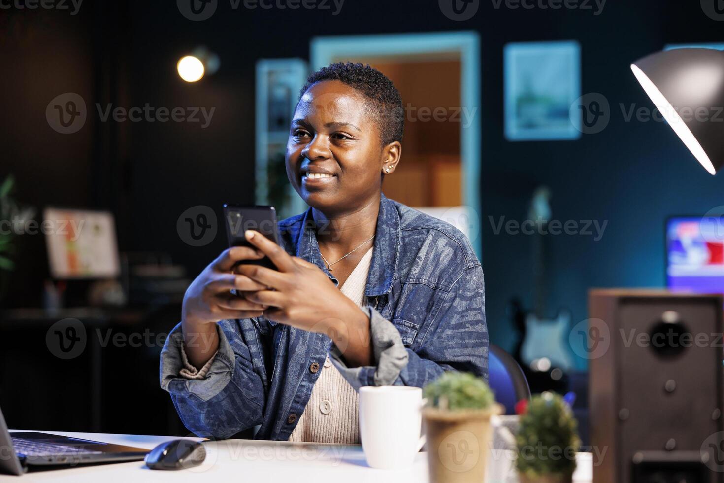 While sitting at her home desk, african american self employed individual checks cell phone for messages. A young black woman using mobile device to chat on social media. photo