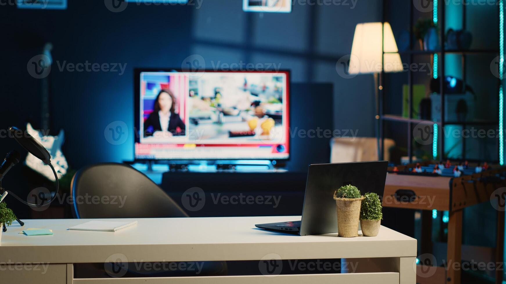 Panning shot of work desk with laptop, microphone and mini house plants in empty home studio interior. Apartment filled with content creation equipment, TV news broadcast running in background photo
