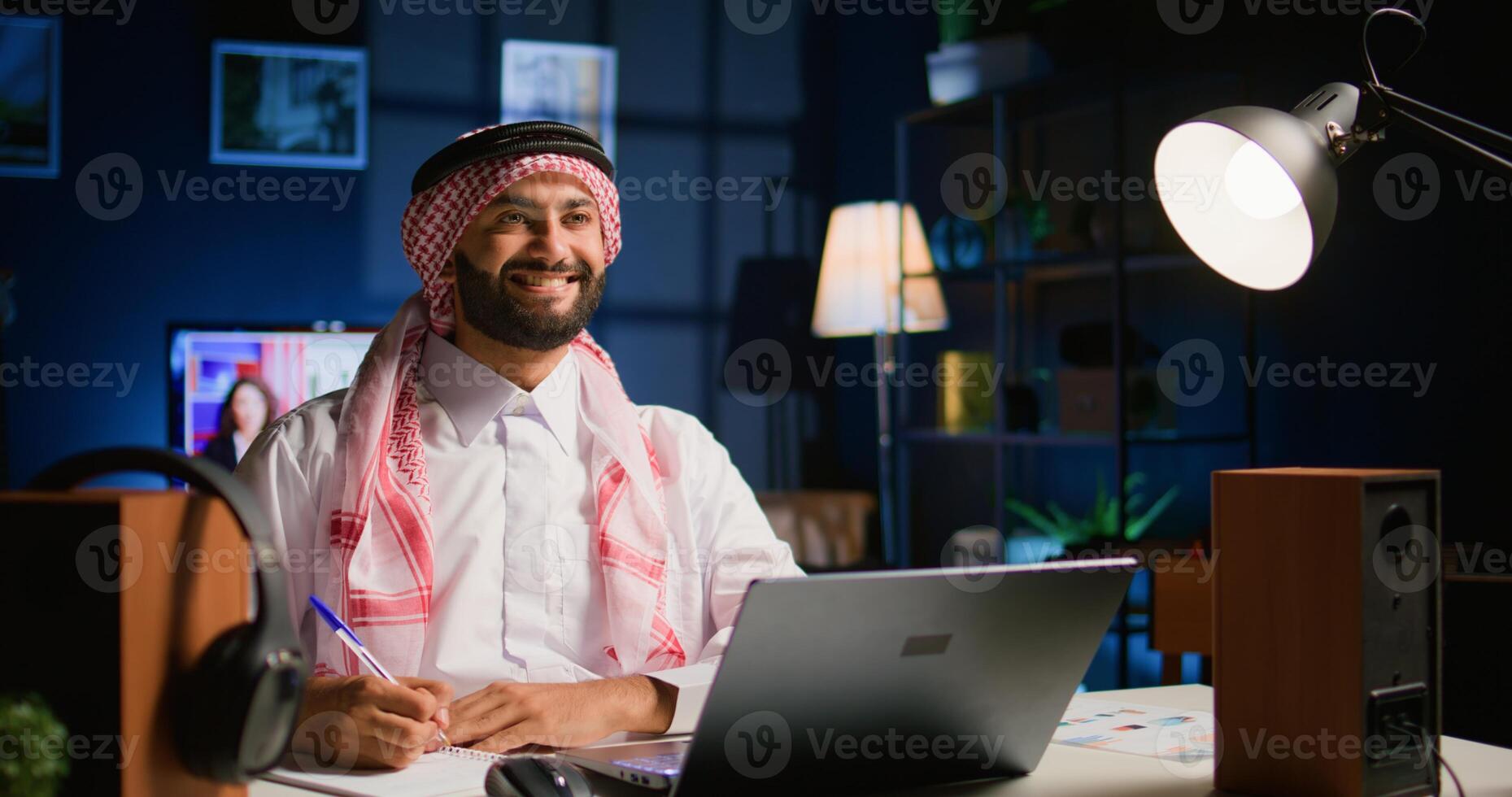 Portrait of happy arab teleworker remotely working in apartment writing on paper. Middle Eastern employee at home cross referencing information, transcribing data from laptop to notepad photo