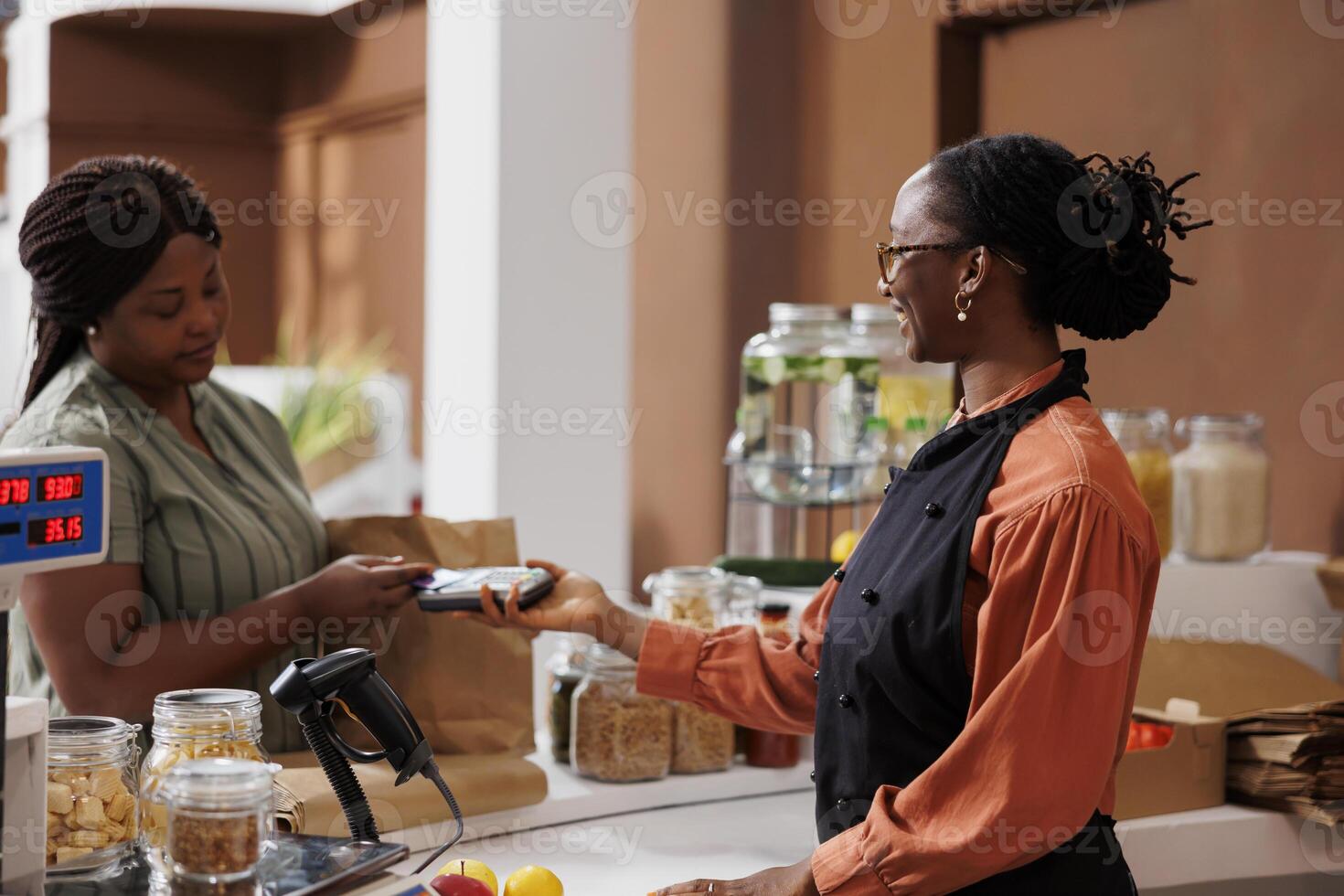 African American woman happily makes cashless payment at market counter using credit card. She smiles as she communicates with the vendor and completes the transaction with a debit and pos machine. photo