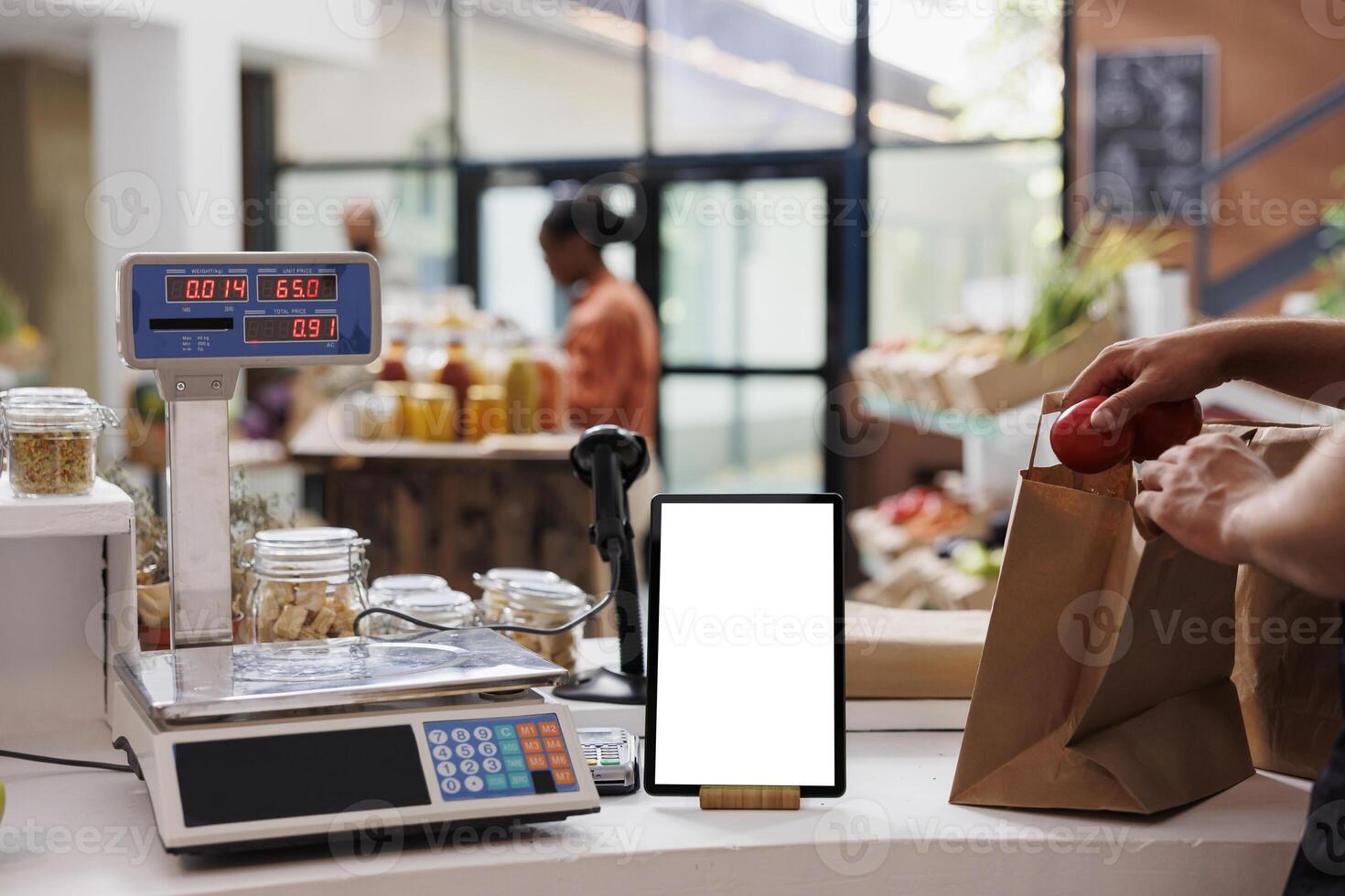 Digital tablet with a white screen display is placed vertically on counter next to a weighing scale. Electronic device showing isolated mockup template while the vendor packs food products in a bag. photo