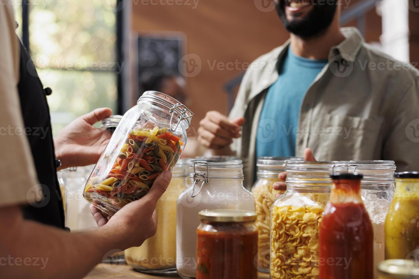 Close-up of a storekeeper holding a glass jar filled with colorful freshly made pasta while a male customer looks on in an environmentally friendly store. photo