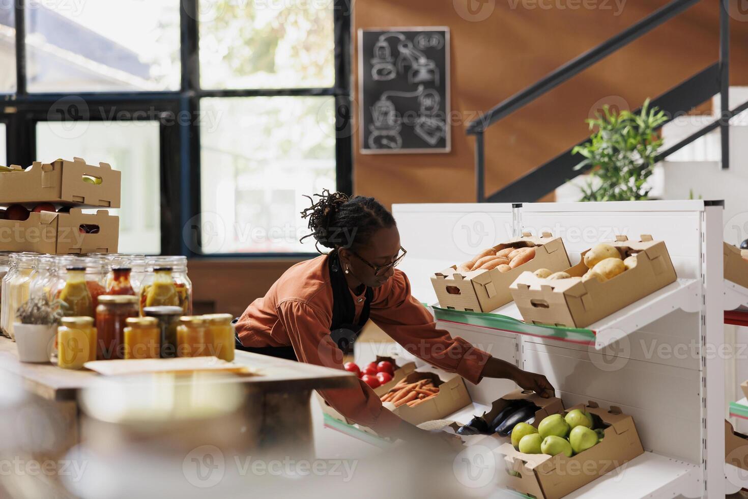 negro mujer con gafas organizando cajas de granja crecido frutas y verduras, preparando para el apertura de cero residuos sostenible almacenar. hembra tendero colocación cajas de bio Produce en estantes. foto