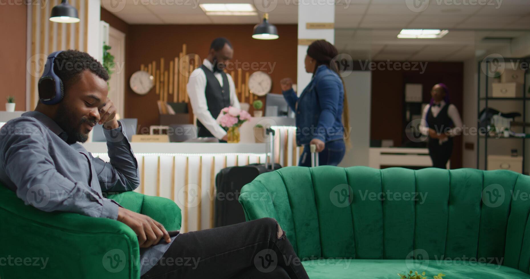 Hotel guest listens music on headset, waiting to check in at front desk and sitting in lounge area on vacation. African american traveller using smartphone to have fun with songs, tourism industry. photo