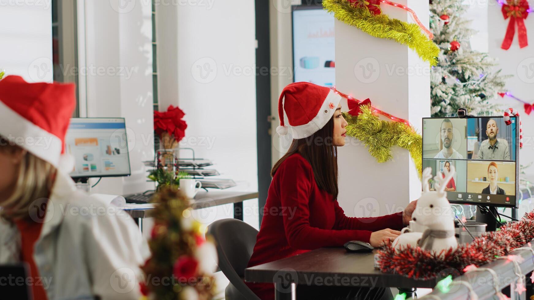 Friendly asian worker in online videocall greeting colleagues during Christmas season in festive decorated office. Employee using webcam for remote teleconference with team in festive adorn workplace photo