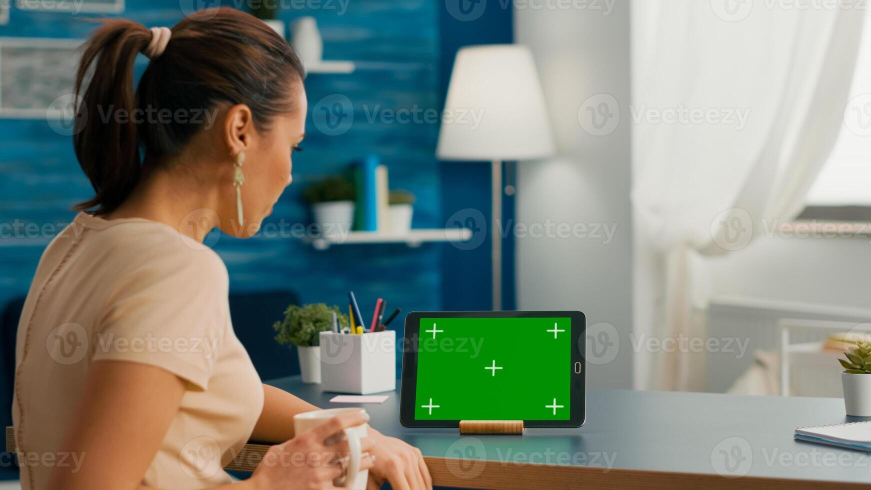 Caucasian female looking at tablet computer with mock up green screen chroma key sitting on office desk in living room. Freelancer woman browsing on social network using isolated device photo