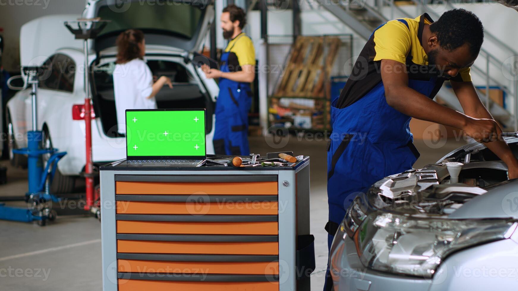 Green screen laptop placed on working bench in busy garage next to professional tools while african american serviceman fixes car. Chroma key device in auto repair shop next to mechanic photo