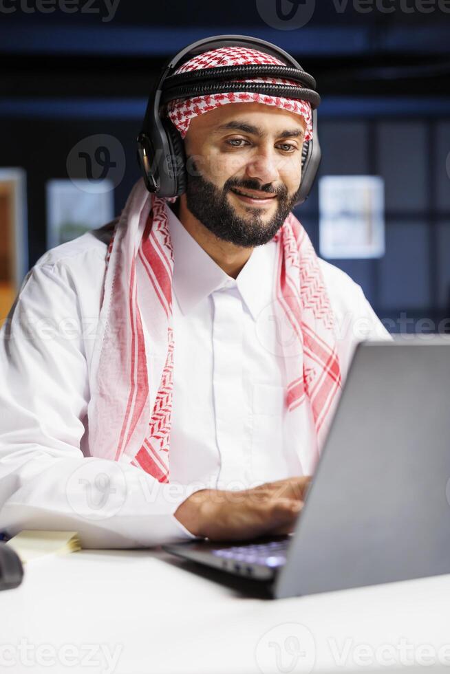 Detailed view of a Muslim entrepreneur in traditional attire working with wireless headphones for calls and researching on his laptop. Youthful Arab man using modern technology. photo