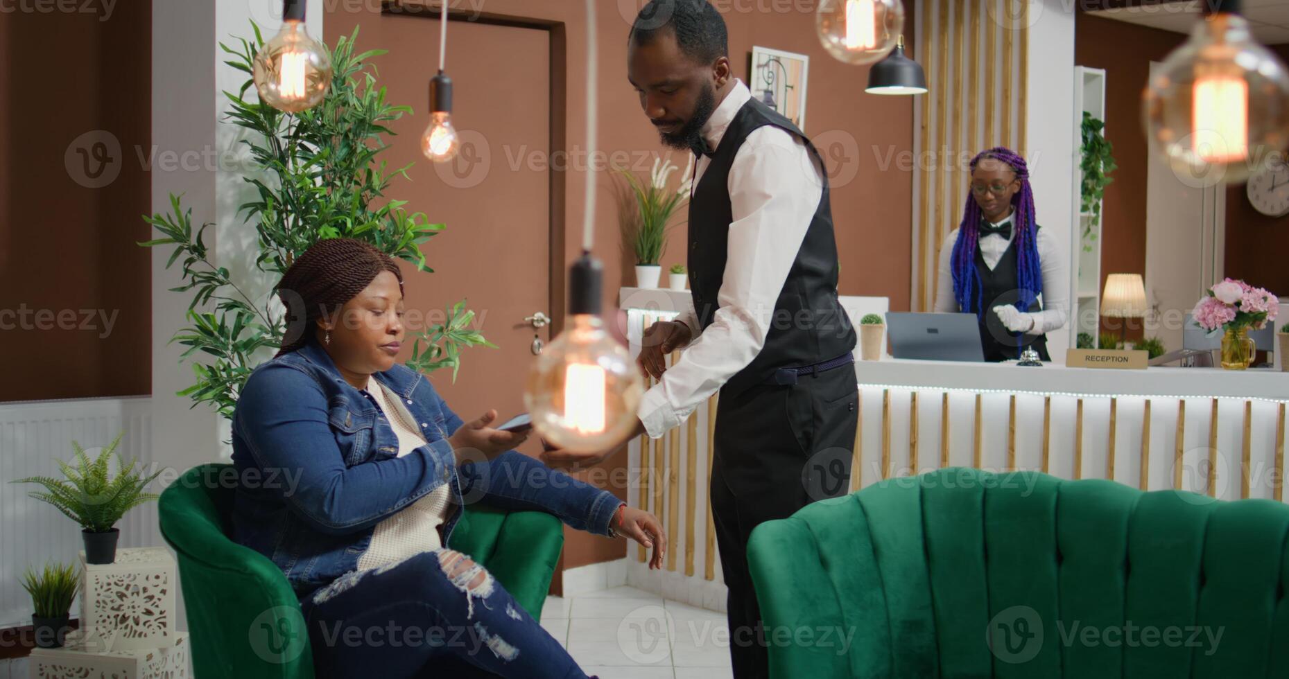 Customer makes nfc payment at pos to pay for coffee cup while waiting on accommodation vacancy in five star hotel. African american traveller talking to bellboy about luxurious service. photo