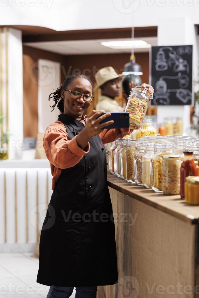 Black shopkeeper promotes organic products and engages customers through social media and calls in bio-food store. She showcases variety of fresh items in glass jars and zero-waste packaging. photo