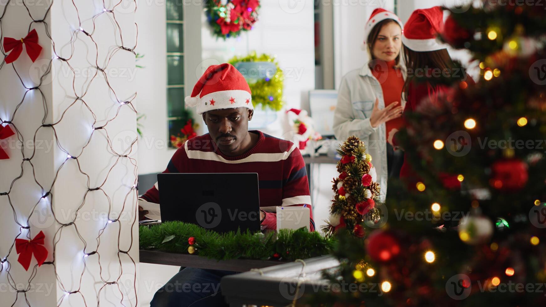African american man filling paperwork in Christmas decorated office, perturbed by disturbing staff members discussing in background. Employee frustrated by colleagues talking too loud photo