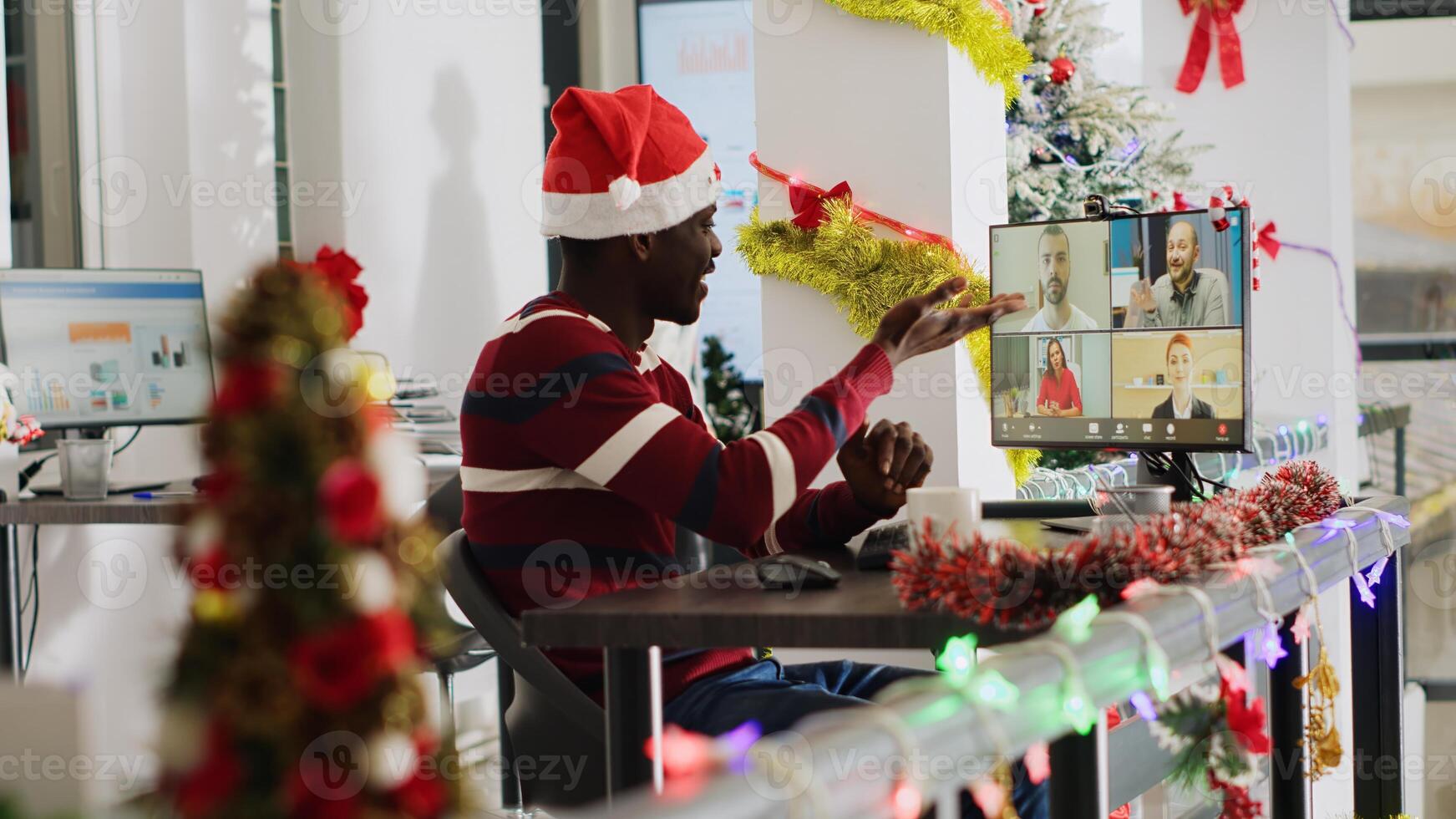 African american worker in online videocall listening to colleagues Christmas decorated office, handheld camera. Employee in remote teleconference receiving feedback from management photo