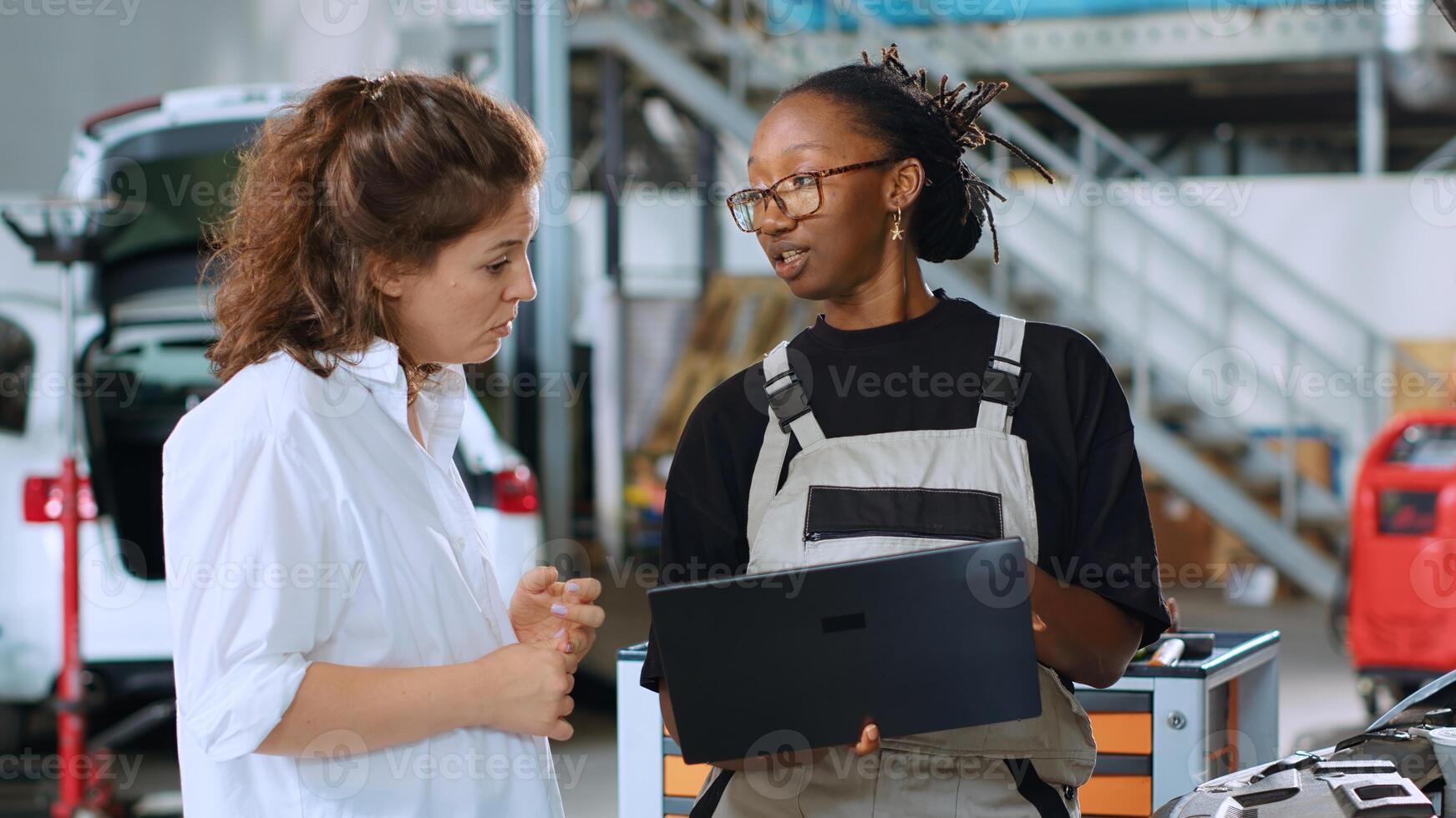Meticulous technician in car service using laptop to calculate repairments invoice for client after fixing damages. Trained expert using device to inform woman of final costs after servicing vehicle photo