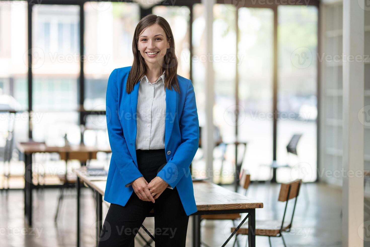 A professional woman with a confident smile stands in a bright, airy office space, dressed in a smart blue blazer with arms crossed. photo
