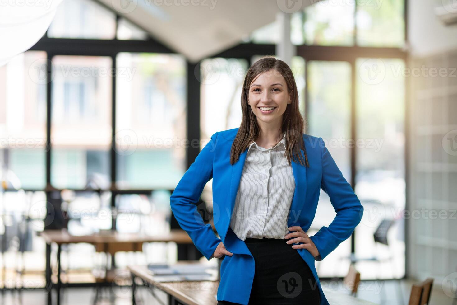 A professional woman with a confident smile stands in a bright, airy office space, dressed in a smart blue blazer with arms crossed. photo