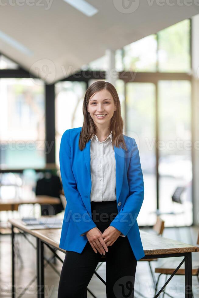 A professional woman with a confident smile stands in a bright, airy office space, dressed in a smart blue blazer with arms crossed. photo