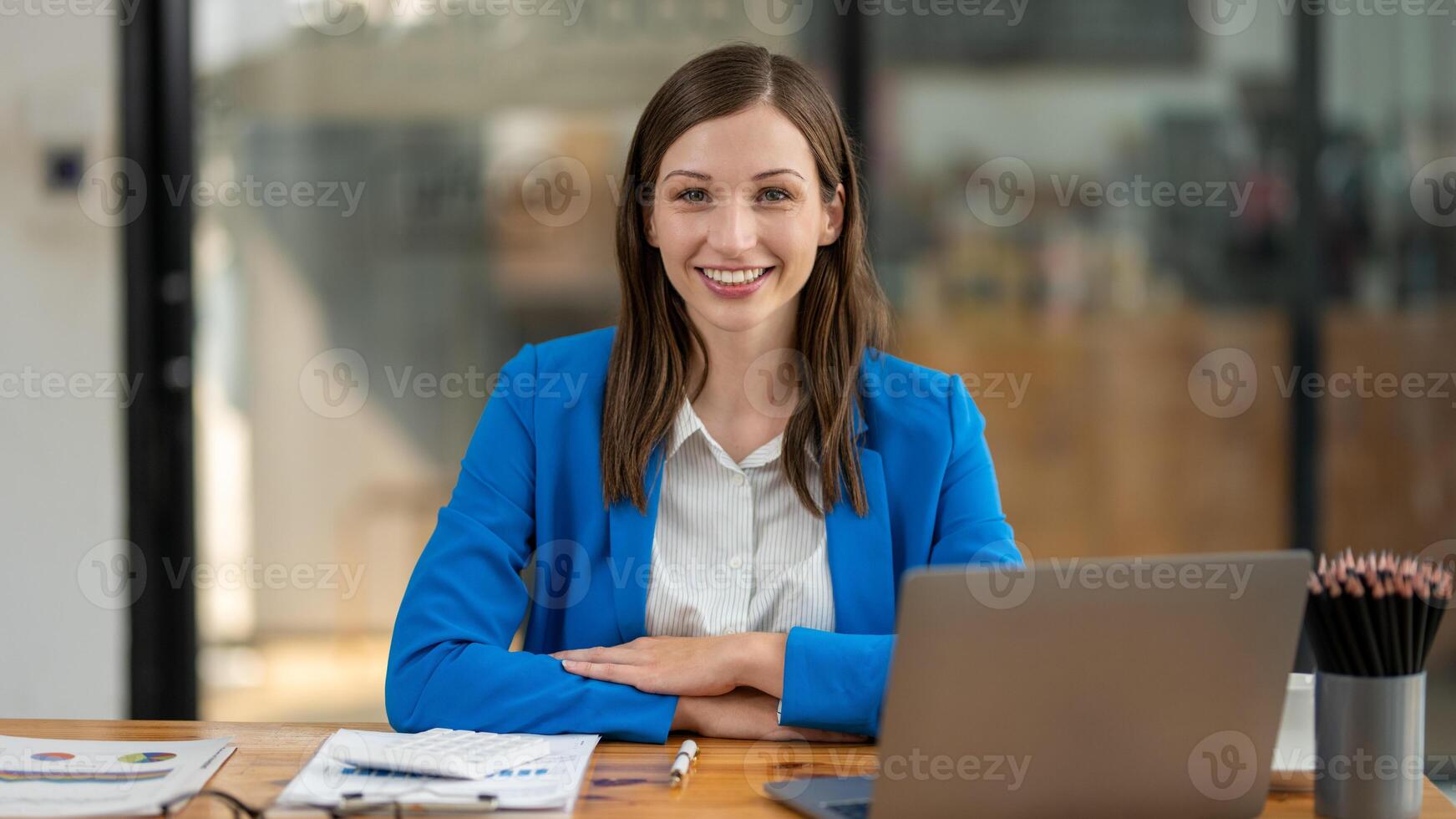 A young, cheerful professional woman sitting at her desk in a bright office environment, looking confidently at the camera. photo