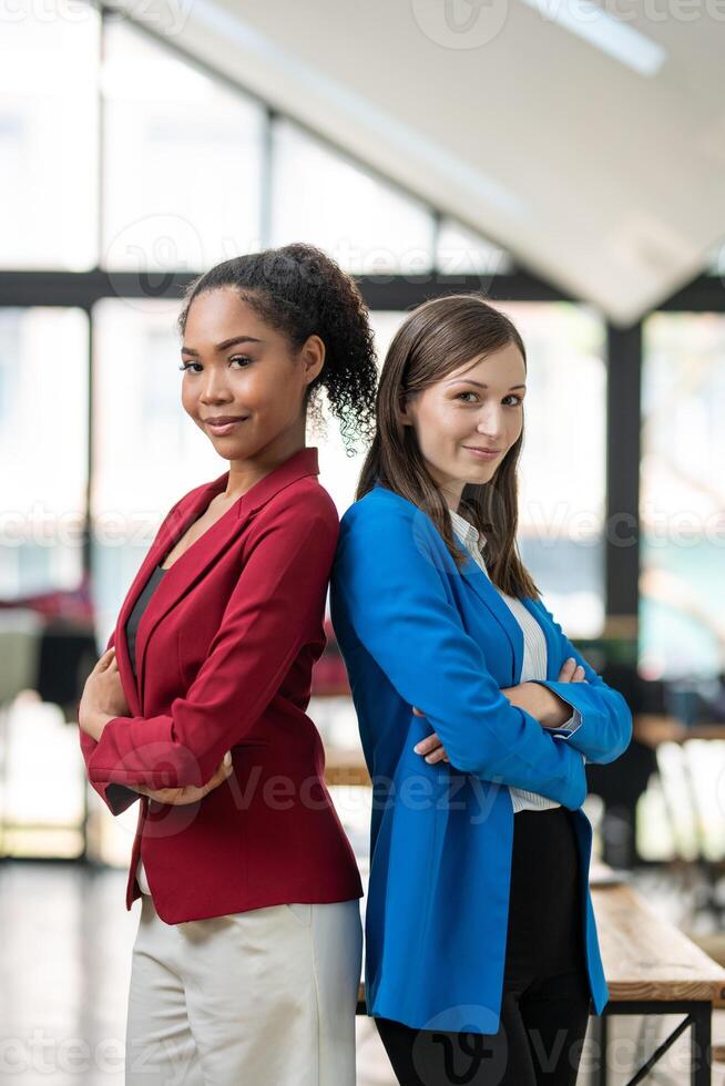 Two diverse businesswomen standing back-to-back, projecting confidence and partnership in a modern office space. photo