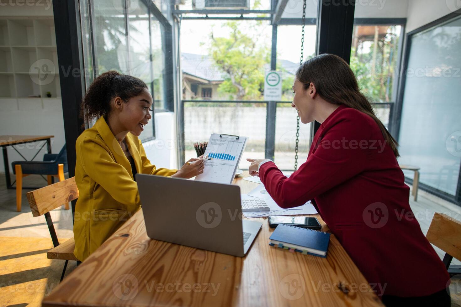 A businesswoman in a yellow blazer engaging in a productive discussion with a colleague in a red jacket over work documents in an office. photo