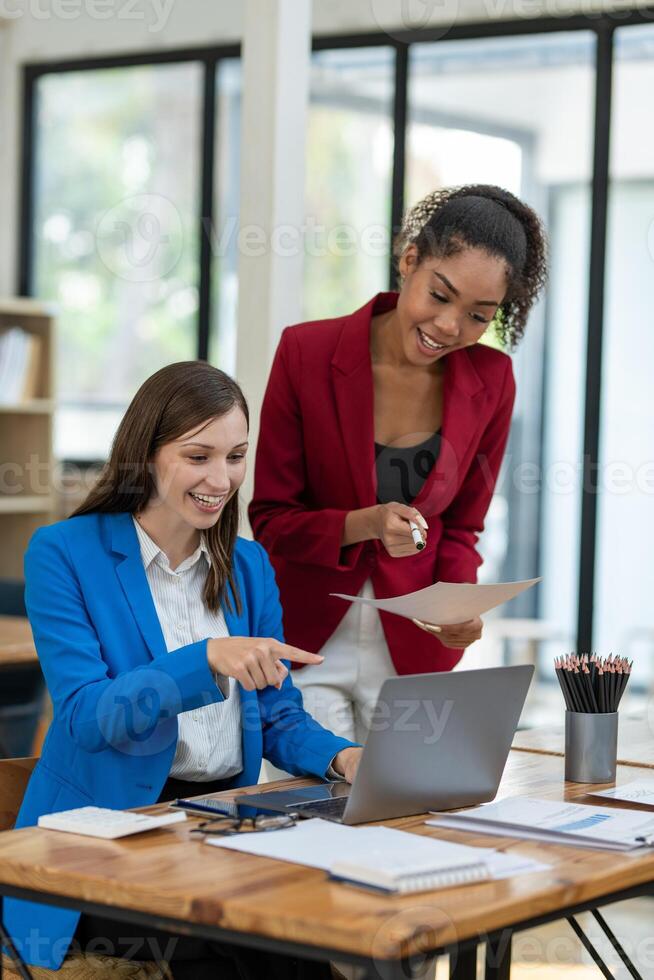 Two professional women in a lively discussion at an office desk, with one in a yellow blazer gesturing and the other showing surprise. photo