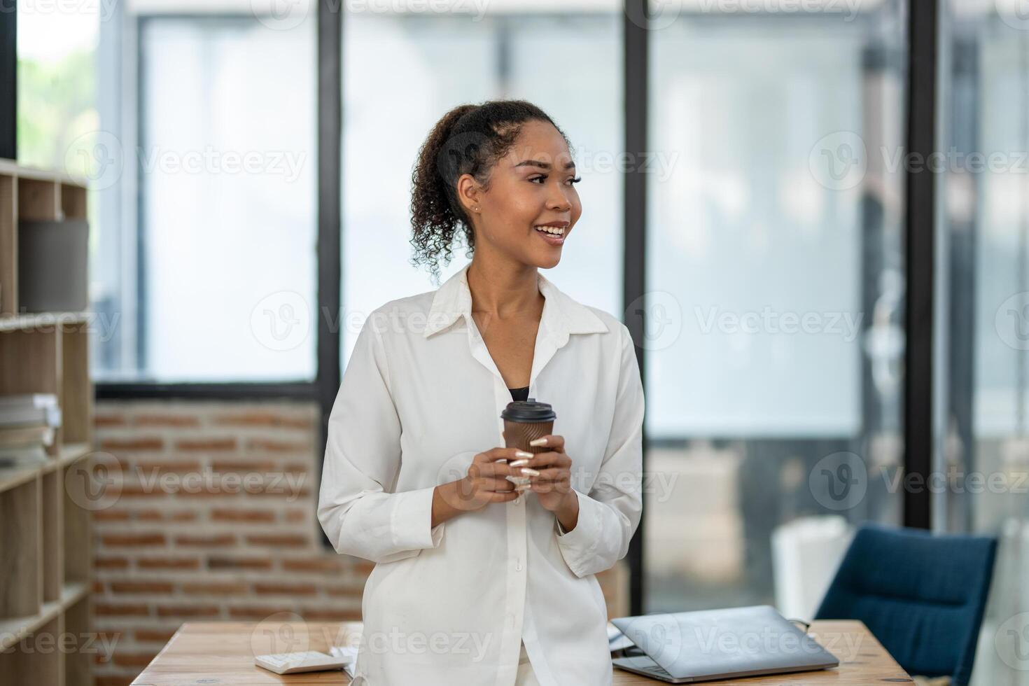 Content young female professional holding a coffee cup during a relaxing break in a modern office setting, looking away with a smile. photo