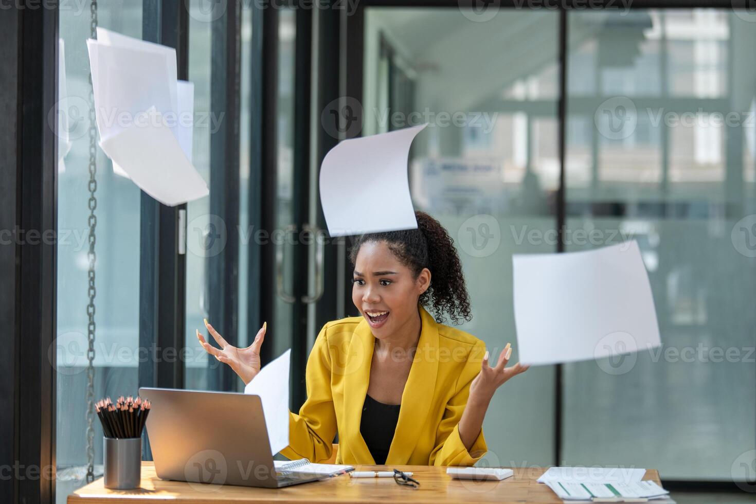 A businesswoman in a yellow jacket expressing frustration and throwing papers into the air at her office workspace. photo