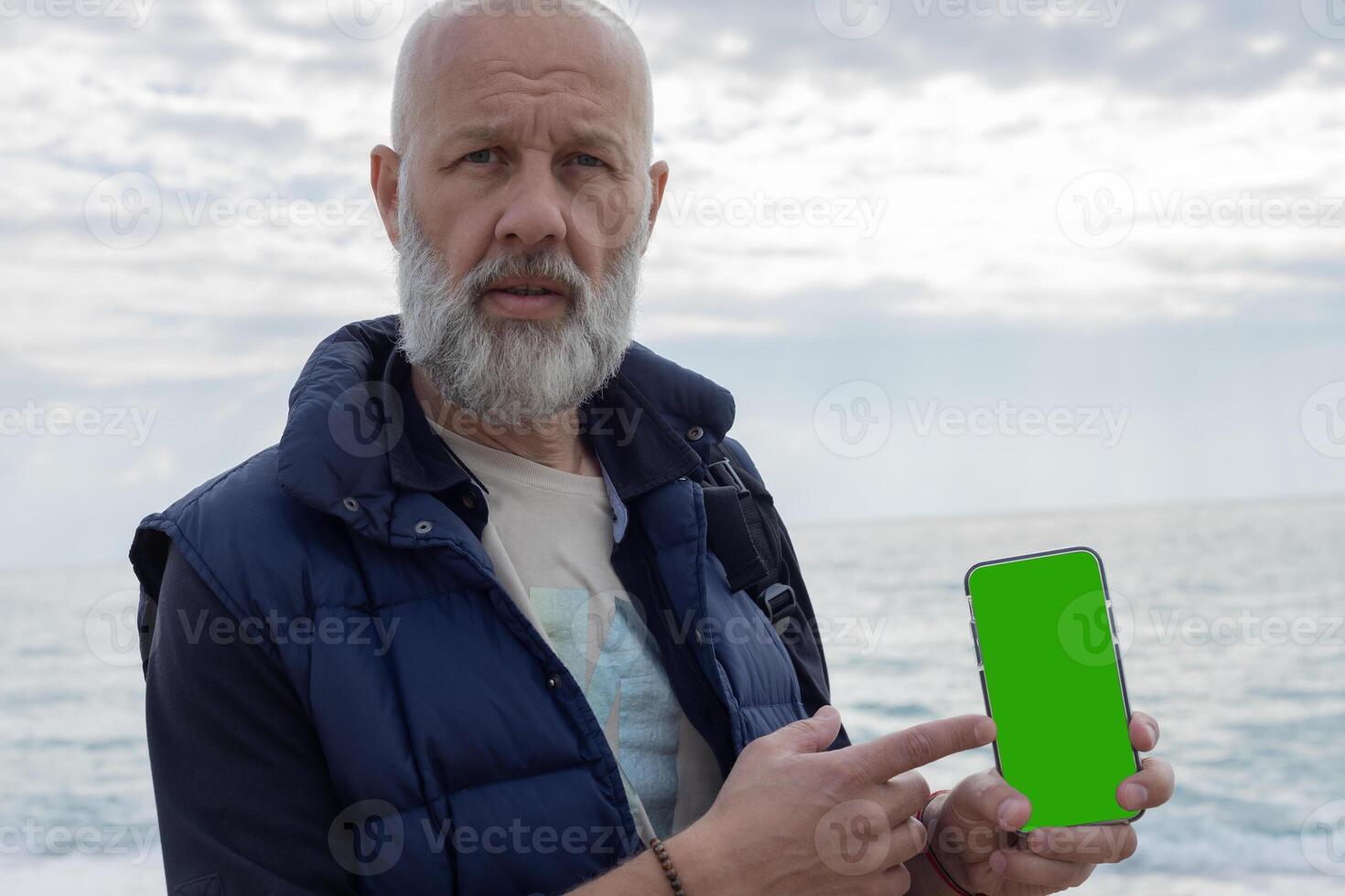 Mockup image of a grey-haired bearded man pointing his finger at a mobile phone with a blank green screen. High quality photo