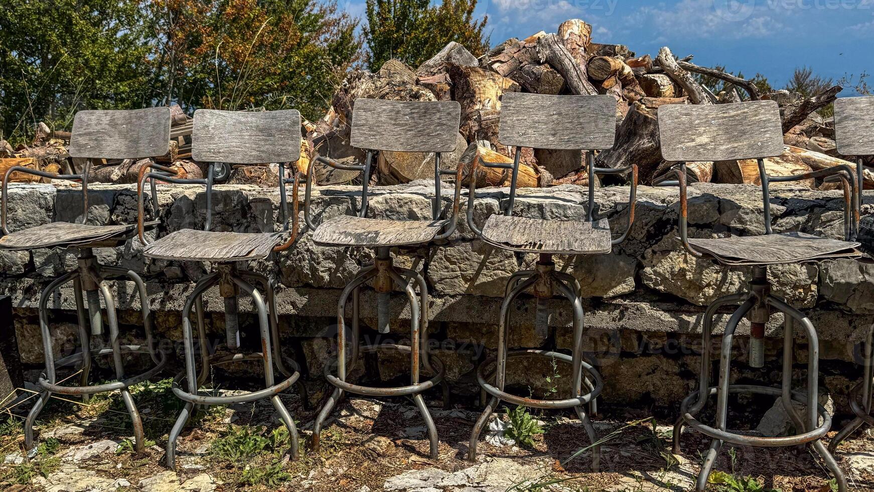 A close-up of six wooden bar stools with rusty metal frames, lined up against a stone wall, against a background of chopped wood and green trees under a clear sky.decay and outdoor decor themes. photo