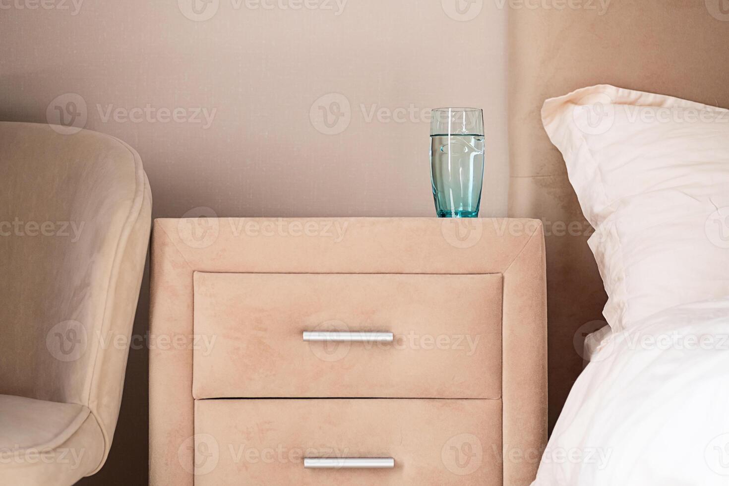 Glass of water on beige bedside table next to bed and chair. photo