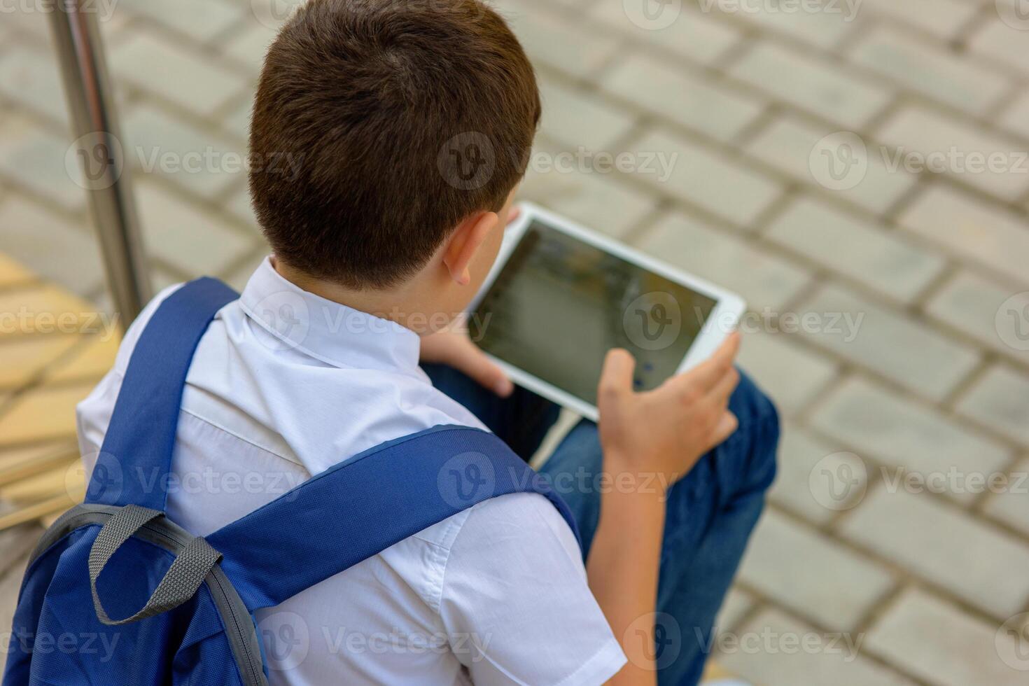 A schoolboy with a blue knapsack sits on the stairs and plays a tablet. Back view photo