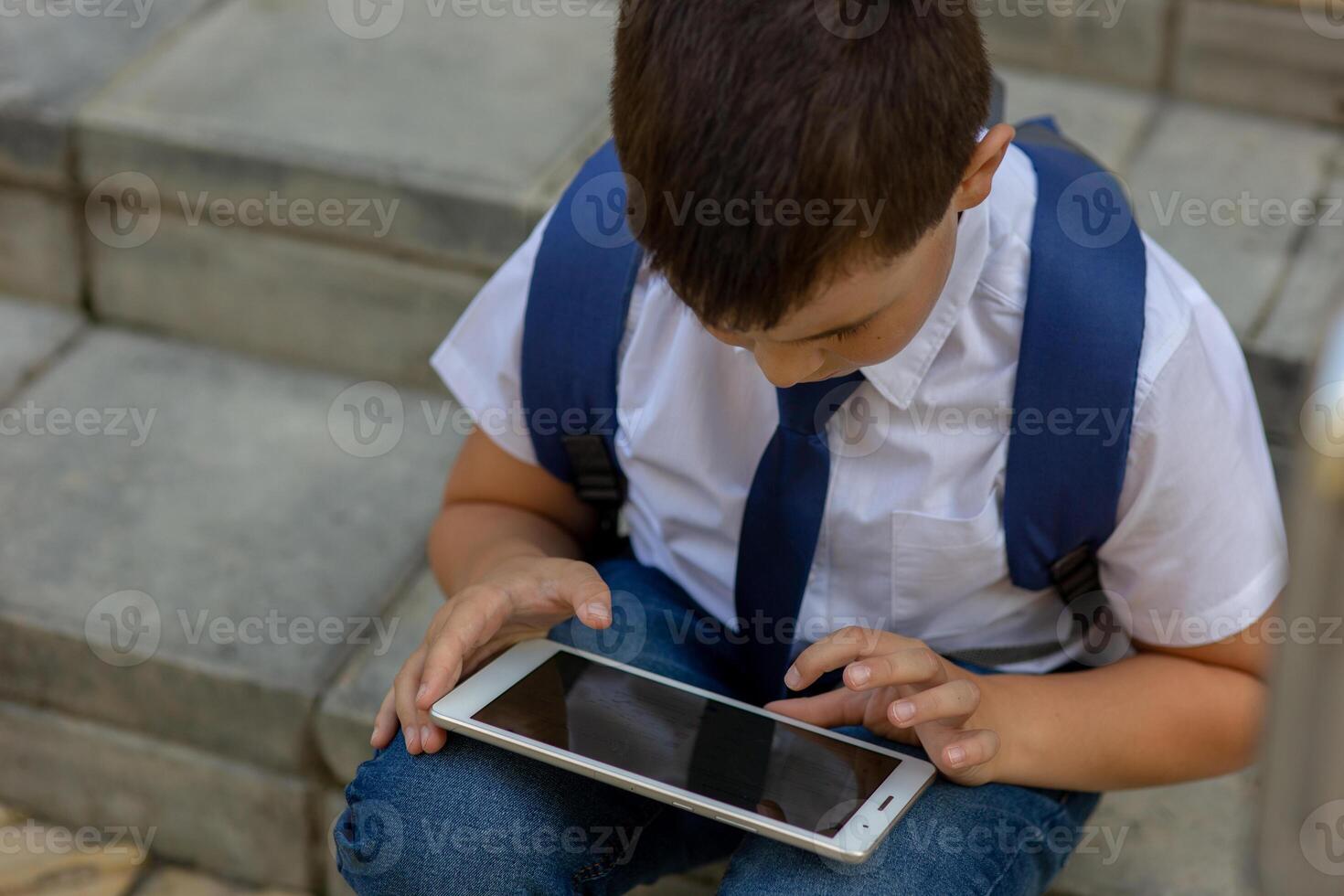 Schoolboy in a white shirt with a blue jacket sits on the stairs and plays with a tablet. photo
