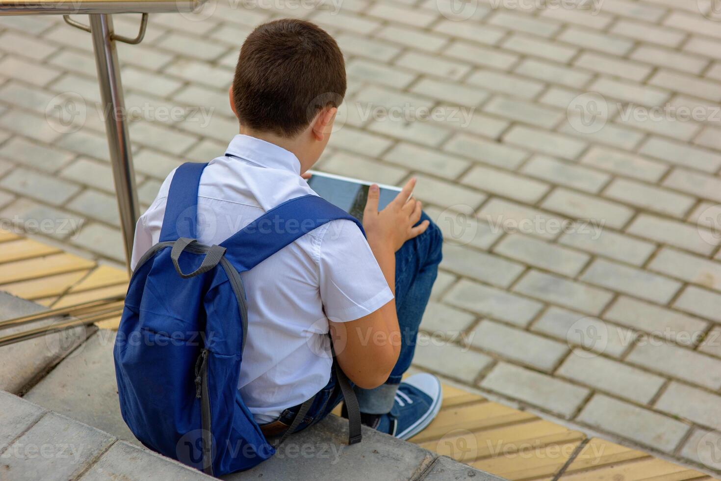 A schoolboy with a blue knapsack sits on the stairs and plays a tablet. Top view photo