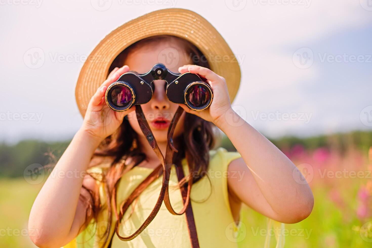 Surprised cute girl 4-5 years old in a straw hat looks through binoculars. photo