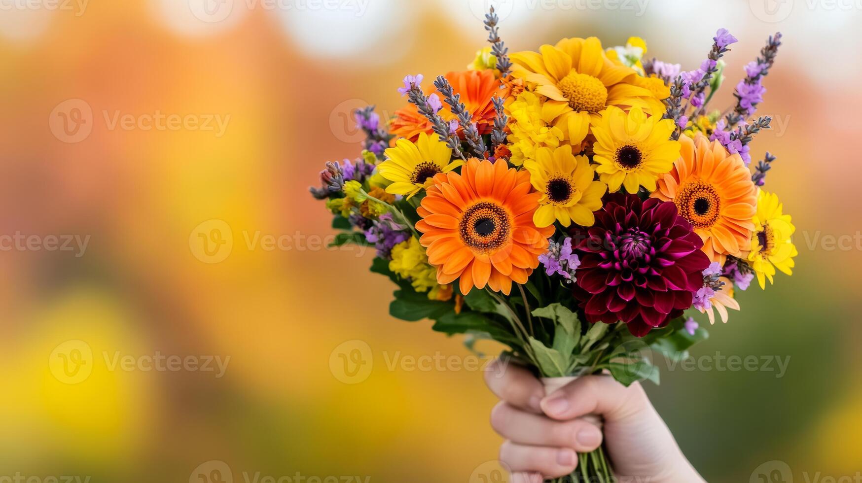 Close up of a hand holding an autumn bouquet with orange and yellow daisies deep red dahlias and sprigs of lavender against a background of soft blurred fall colors photo