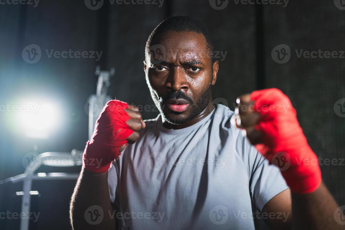 Boxer in gym. Aggressive African man fighter resting after training boxing ready for fight looking at camera. Strong sweated man training punches looking concentrated straight preparing for sparring photo